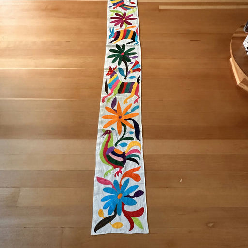Colorful embroidered table runner featuring vibrant flowers and stylized animals, laid out on a wooden floor.