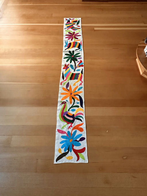 Colorful embroidered table runner featuring vibrant flowers and stylized animals, laid out on a wooden floor.