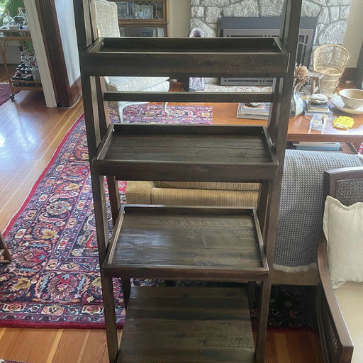 Wooden shelving unit with five open shelves in a cozy living room setting, featuring a patterned rug, a stone fireplace, and surrounding furniture.