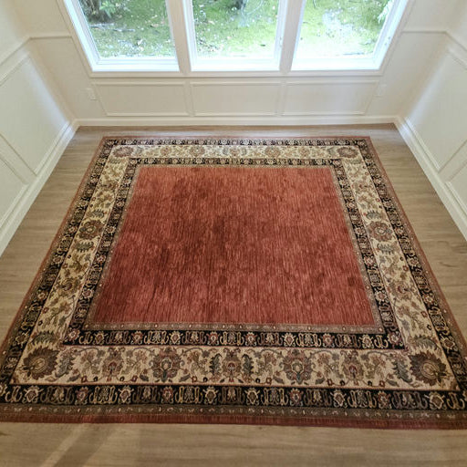 A large red area rug with intricate patterned borders sits on a wooden floor, surrounded by light wooden paneling and large windows.