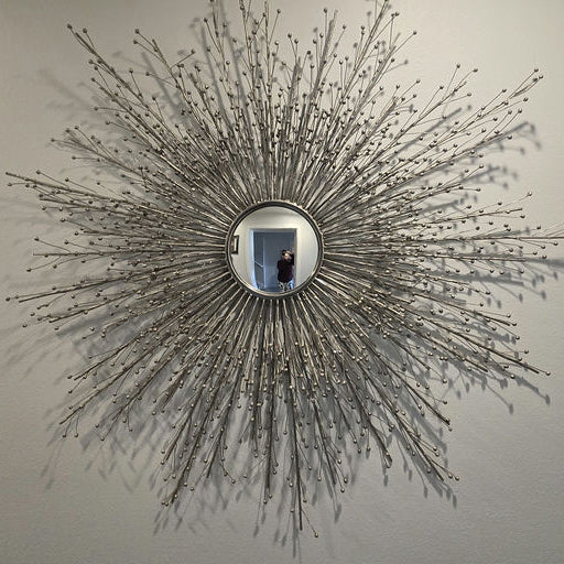 A decorative sunburst mirror with a silver frame and protruding branches, reflecting a person standing in front of it against a light wall.