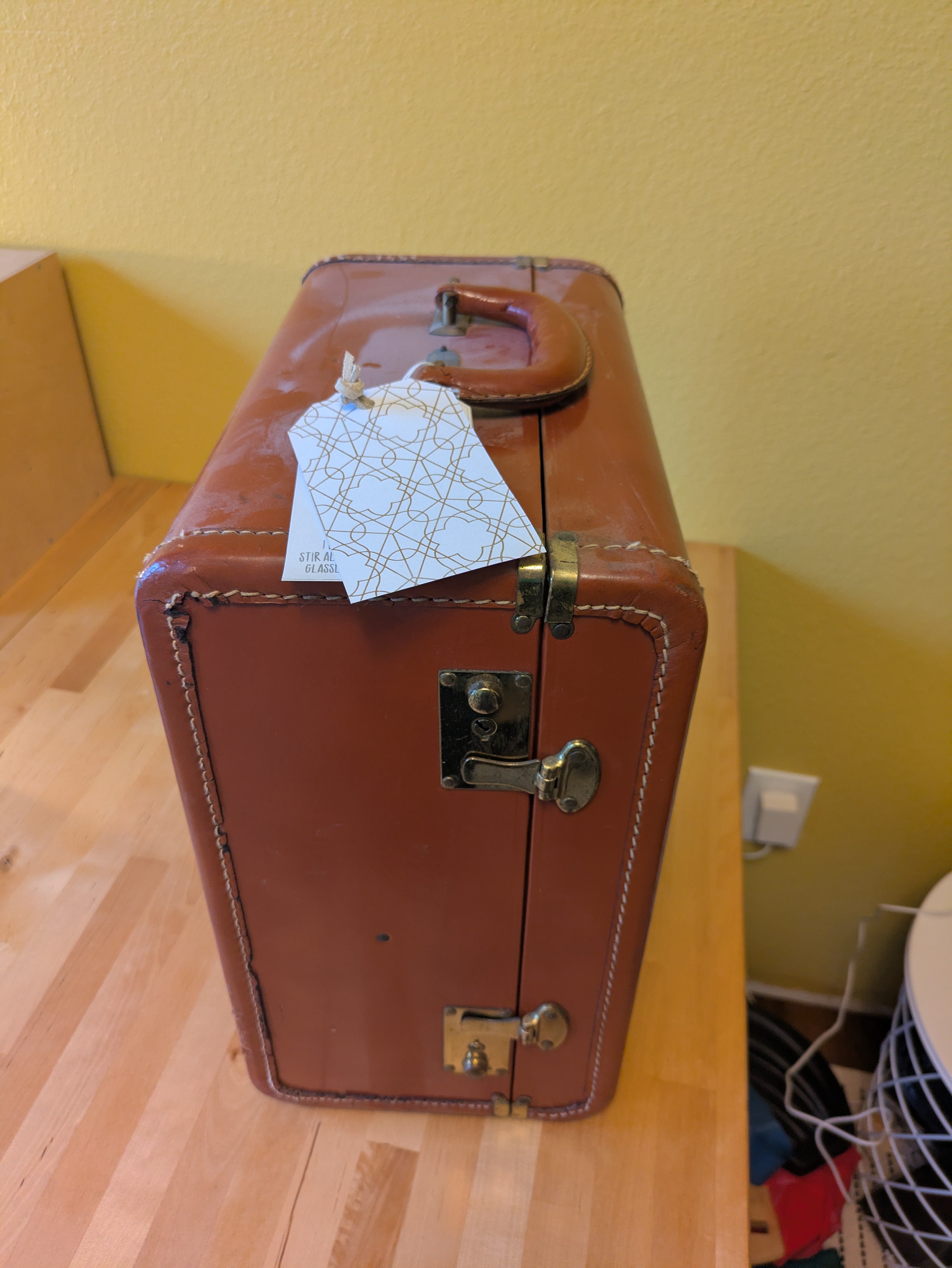 A vintage brown suitcase with a handle, sitting on a wooden surface. It has a tag attached, featuring a geometric pattern and text.