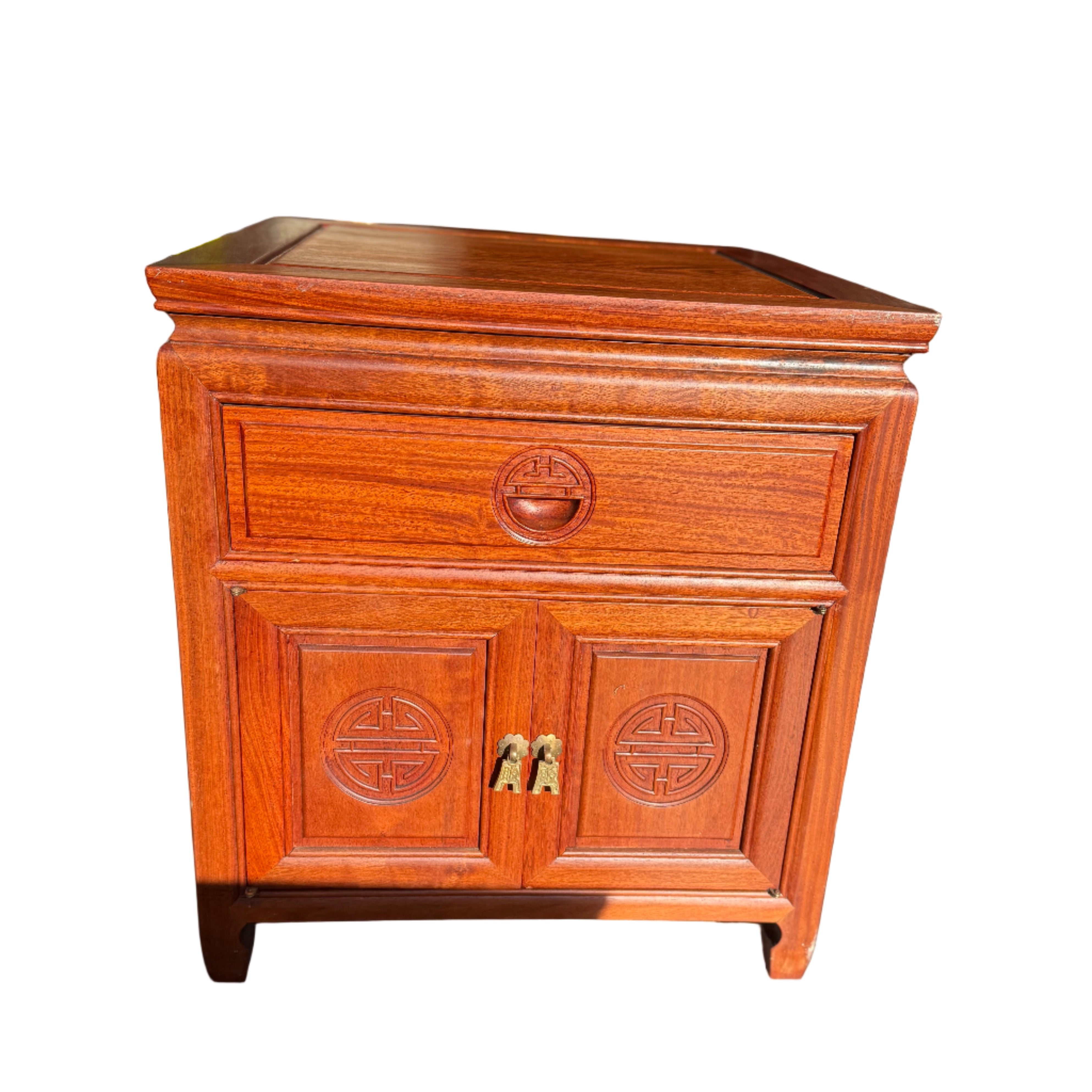 A wooden nightstand with intricate carvings and a polished finish sits in a dimly lit room, casting shadows on the floor. A lamp is visible in the background.