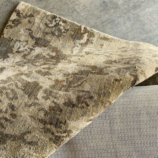 A patterned, light-colored rug is partially draped over a textured, neutral fabric surface, showcasing intricate designs against a concrete floor.