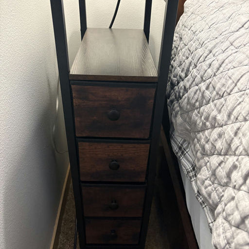 A wooden nightstand with four small drawers and a flat surface on top, housing a lamp. Positioned next to a bed. Wall and carpet visible.