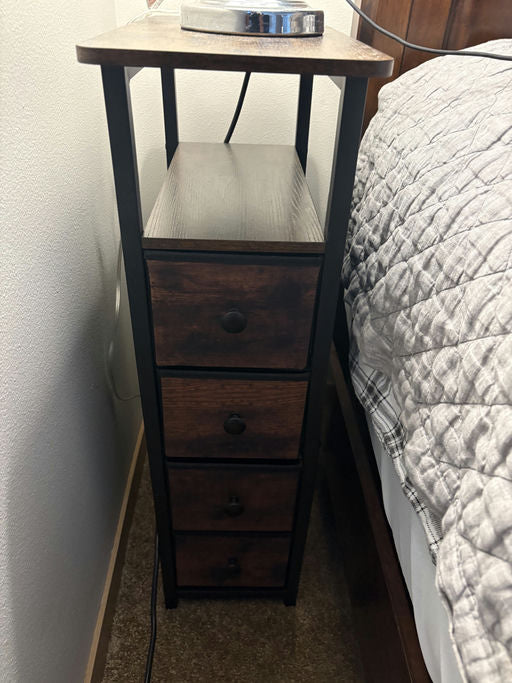 A wooden nightstand with four small drawers and a flat surface on top, housing a lamp. Positioned next to a bed. Wall and carpet visible.