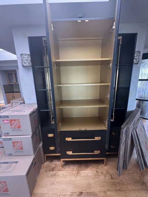 A modern black cabinet with gold interior and shelves stands among cardboard moving boxes and packing materials on a wooden floor.