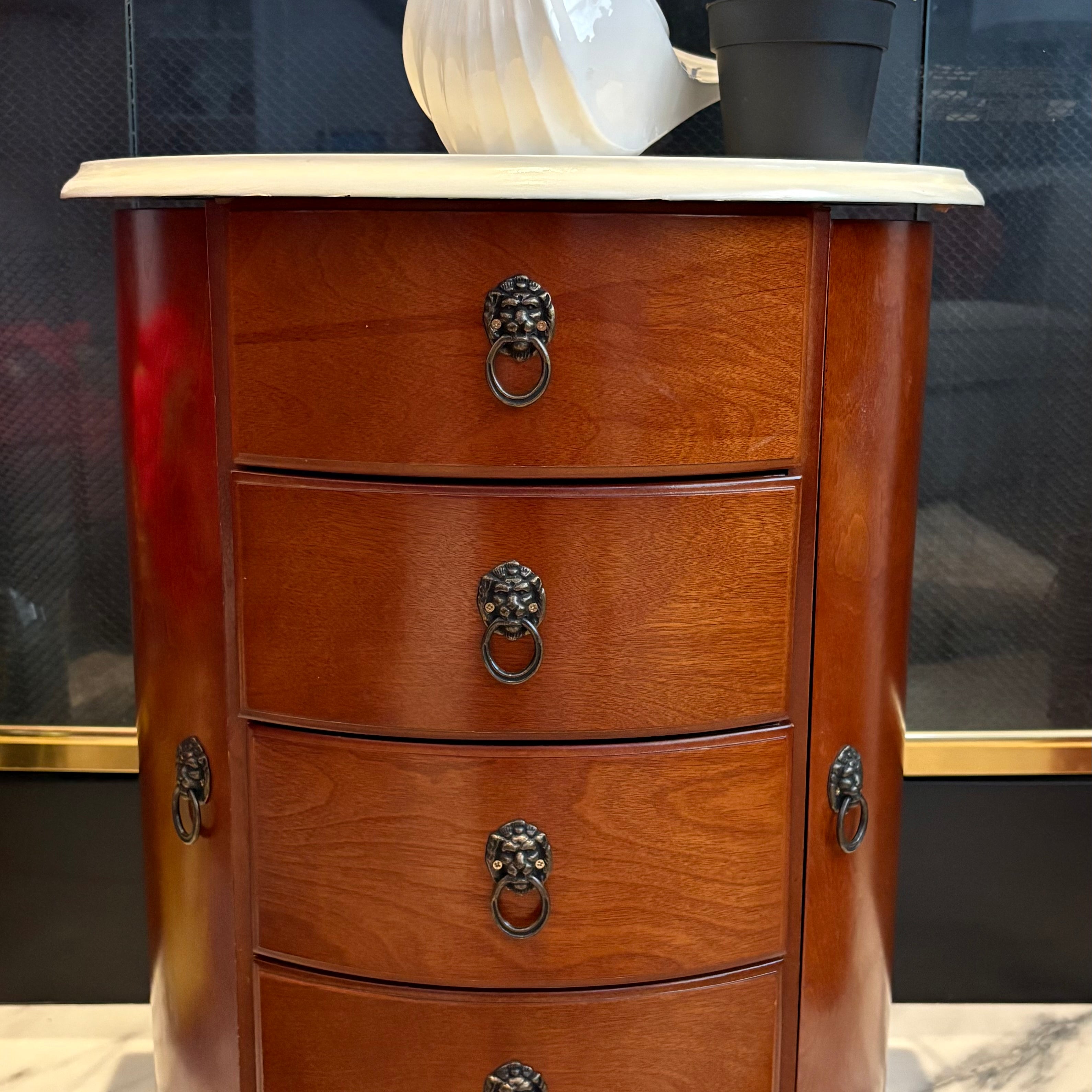A round wooden cabinet with five drawers, featuring lion's head handles. On top, a white decorative object and a potted plant with red flowers.