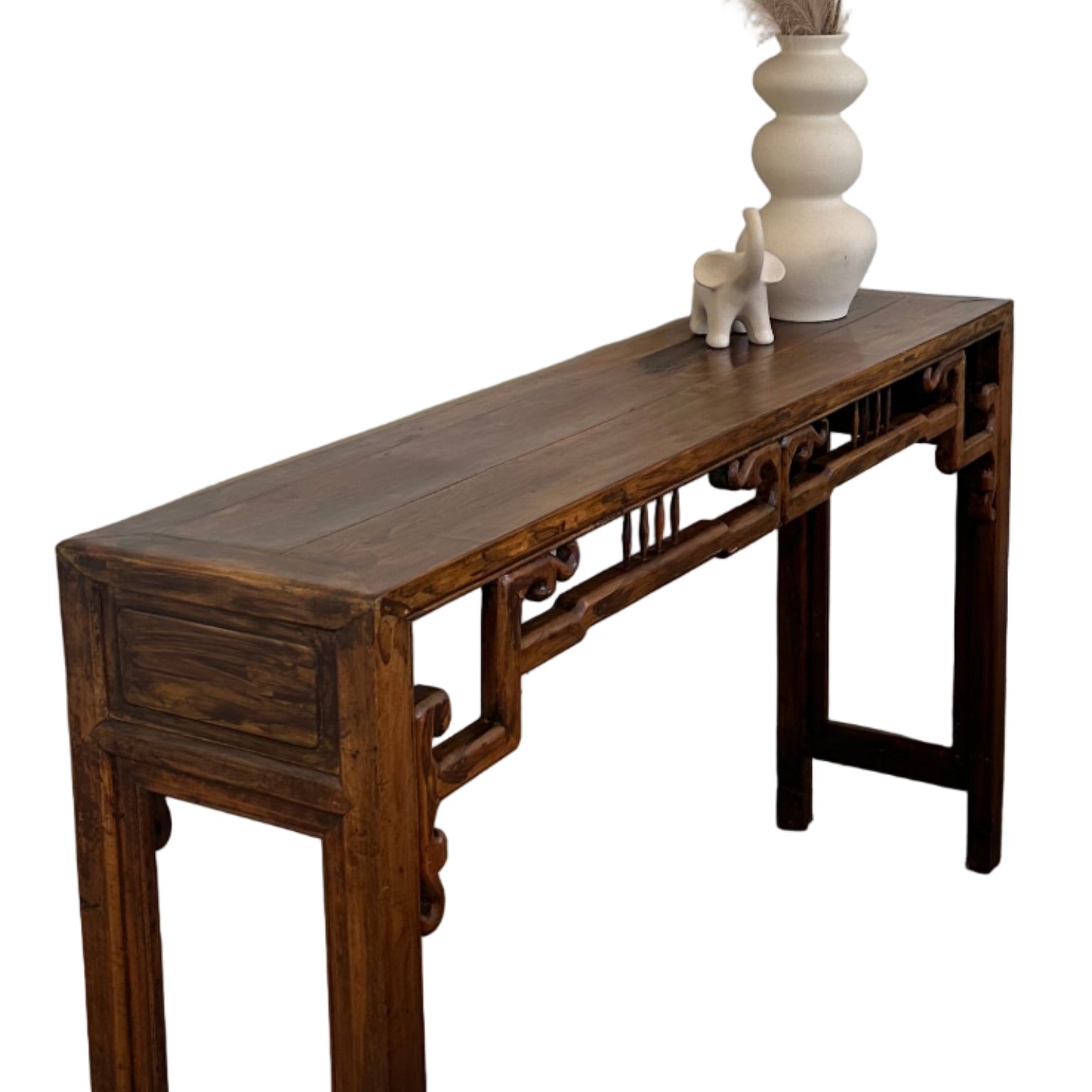 A wooden console table with intricate details stands against a white wall, adorned with a white vase and decorative pampas grass. A gray sofa is nearby.