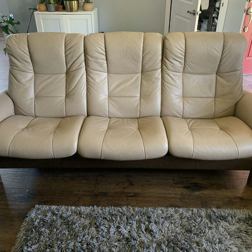 A light beige leather sofa with three seats and plush backing, sitting on a wooden floor with a textured rug in front, and a decorated shelf in the background.