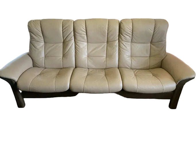 A light beige leather sofa with three seats and plush backing, sitting on a wooden floor with a textured rug in front, and a decorated shelf in the background.