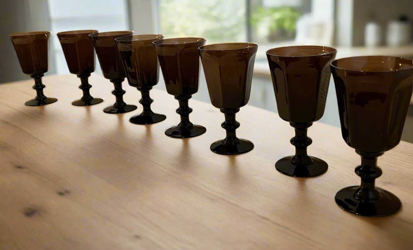 A row of eight dark glass goblets with black stems sits on a white surface under soft natural light, creating a sophisticated display.