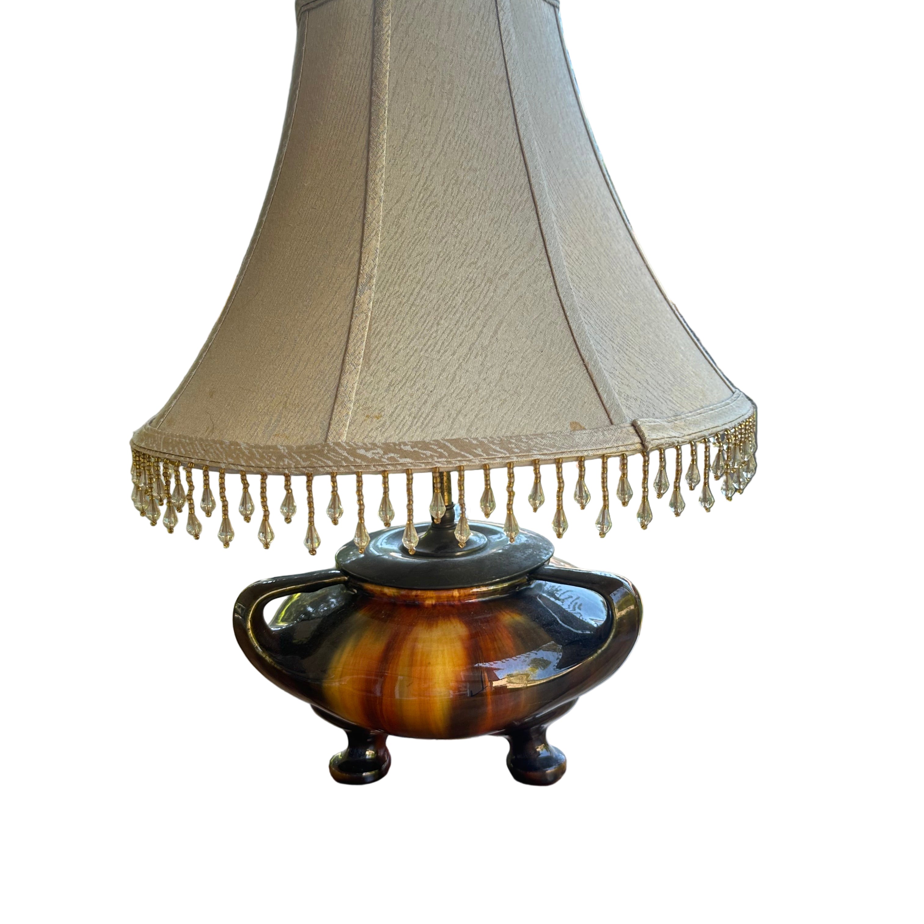 A decorative lamp with a cream lampshade and a colorful, vintage-style base sits on a wooden table, surrounded by greenery and natural light.