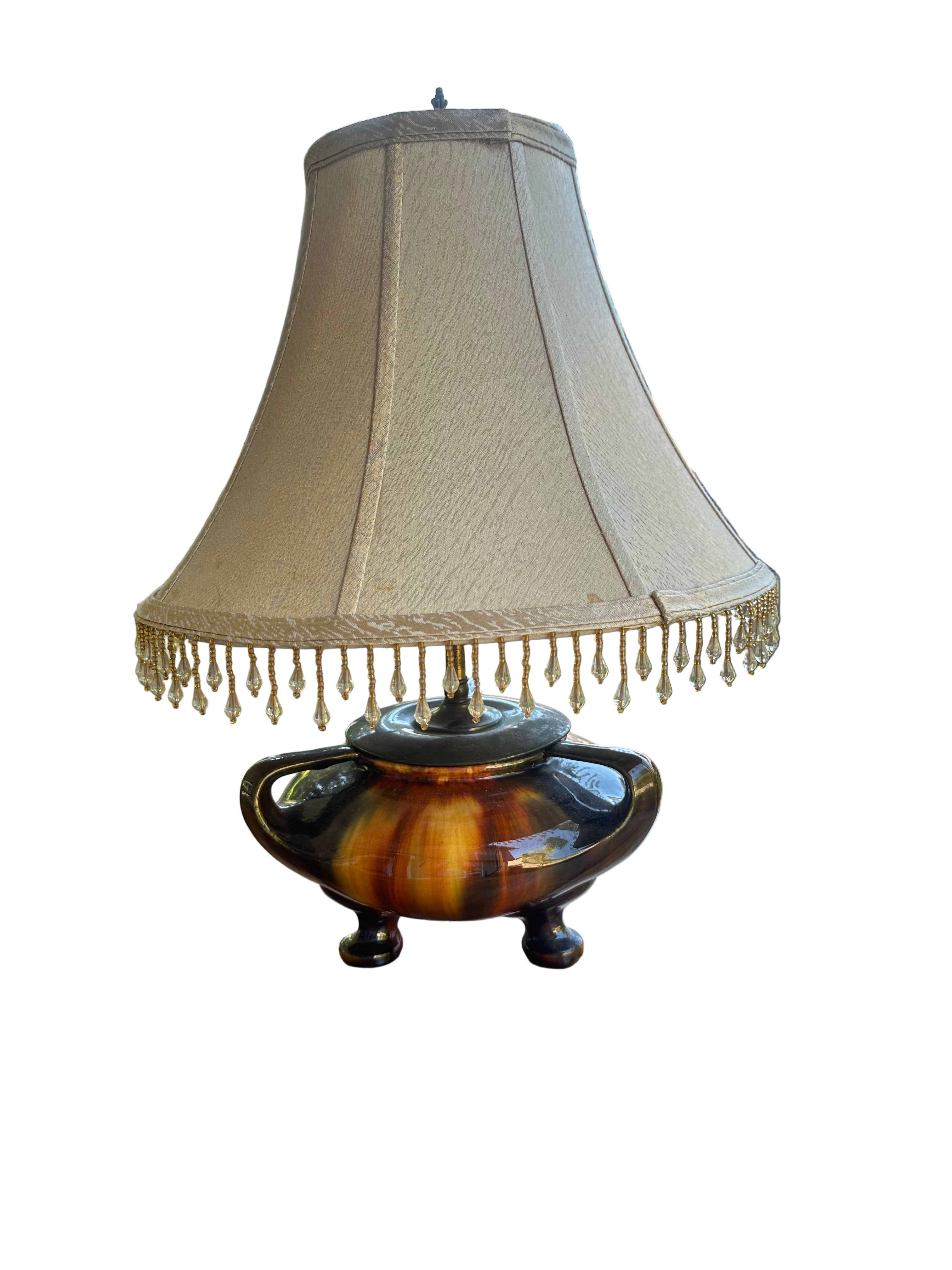 A decorative lamp with a cream lampshade and a colorful, vintage-style base sits on a wooden table, surrounded by greenery and natural light.