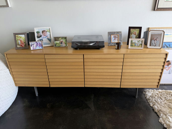 A wooden sideboard displays framed photos of family and friends, accompanied by a turntable and a small candle, against a neutral wall.