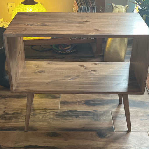 A wooden side table with an open storage space and tapered legs, situated in a cozy room with a plant and decorative lamp in the background.