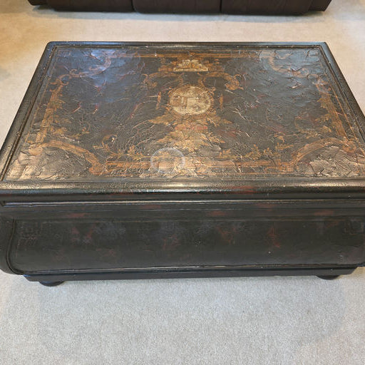 An antique black trunk with intricate artwork on top, featuring ornate patterns and designs. It's placed on a light carpet in a cozy room.