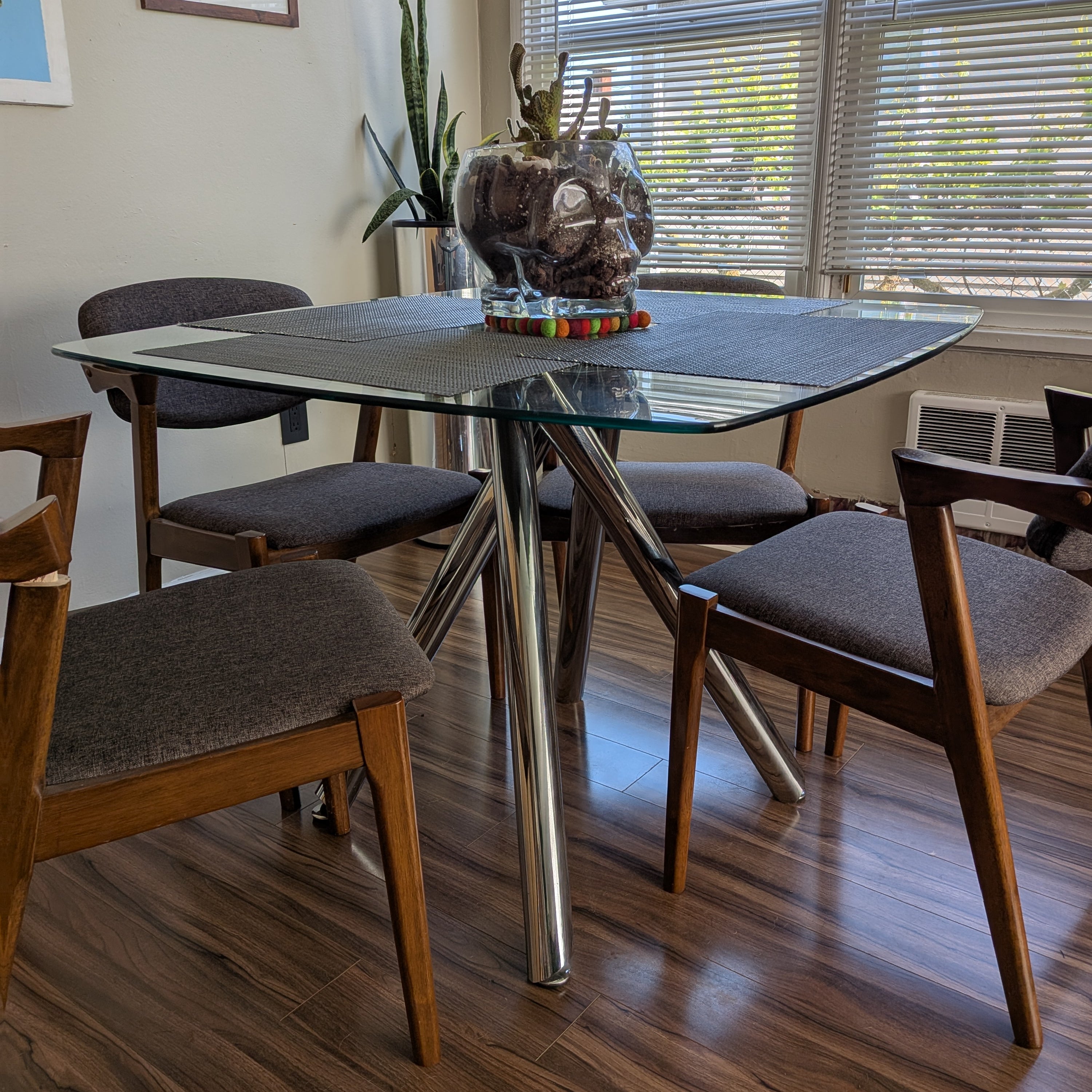A modern dining area with a glass-top table supported by chrome legs, surrounded by wooden chairs. A decorative glass skull holds plants.