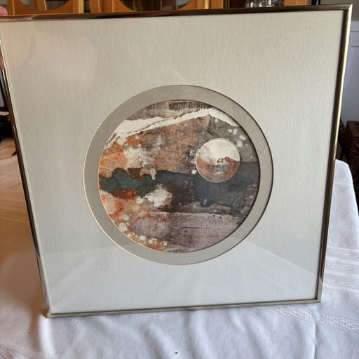 Framed abstract artwork featuring circular designs in earthy tones of orange, gray, and white, displayed on a textured surface.