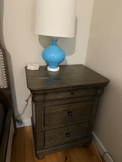 A wooden nightstand with three drawers, topped by a blue lamp with a white shade. A small decorative item sits beside the lamp, on a wooden floor.