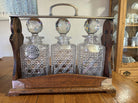 Three crystal decanters labeled "Whisky," "Sherry," and "Gin," elegantly displayed in a wooden holder, set on a wooden table.