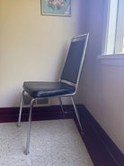 A metal and black vinyl chair is positioned in a corner of a room, beside a window and a floral artwork on the wall. The floor is tiled.