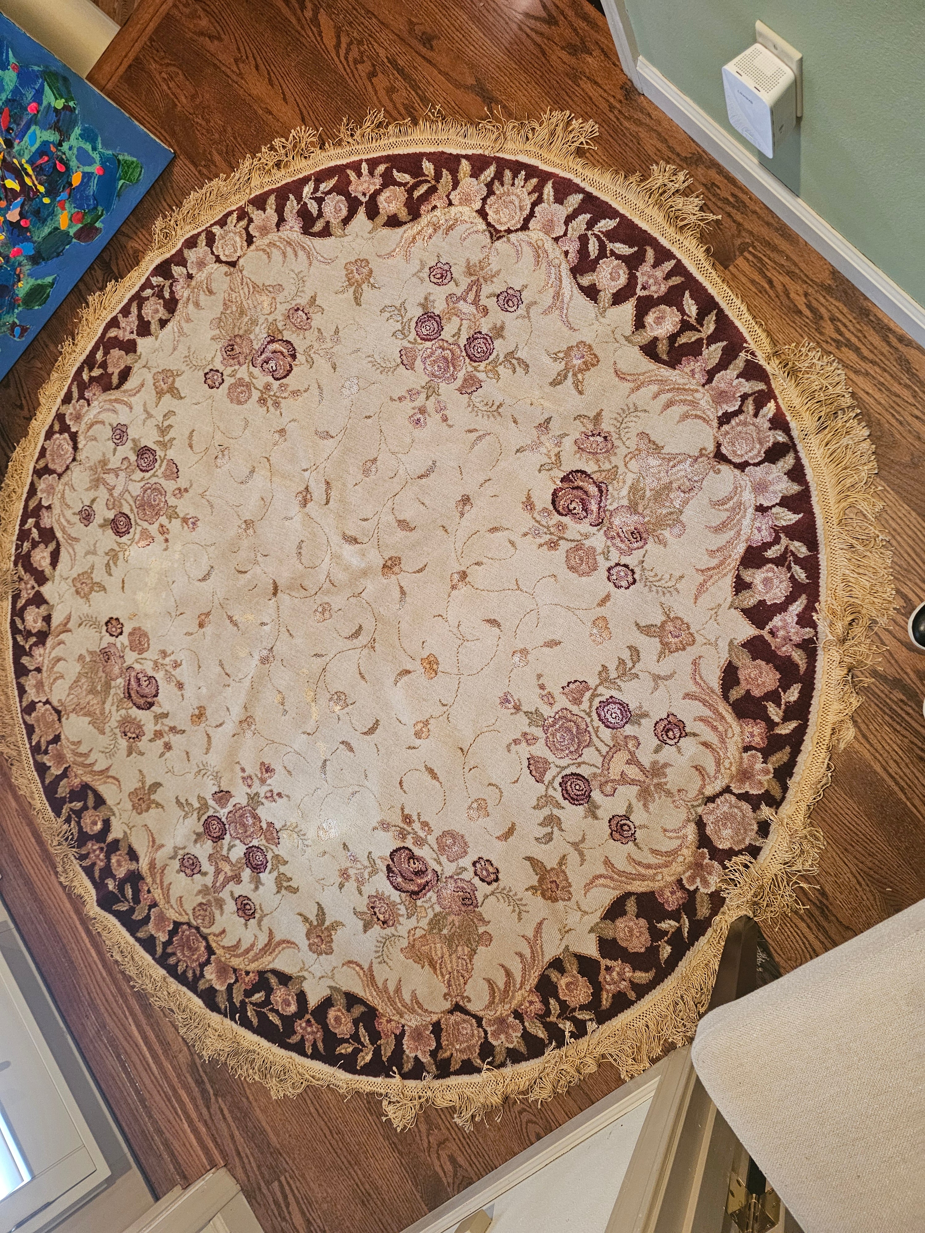 A round, intricately designed area rug with floral patterns in soft shades of cream, pink, and burgundy, adorned with fringed edges, on hardwood floor.