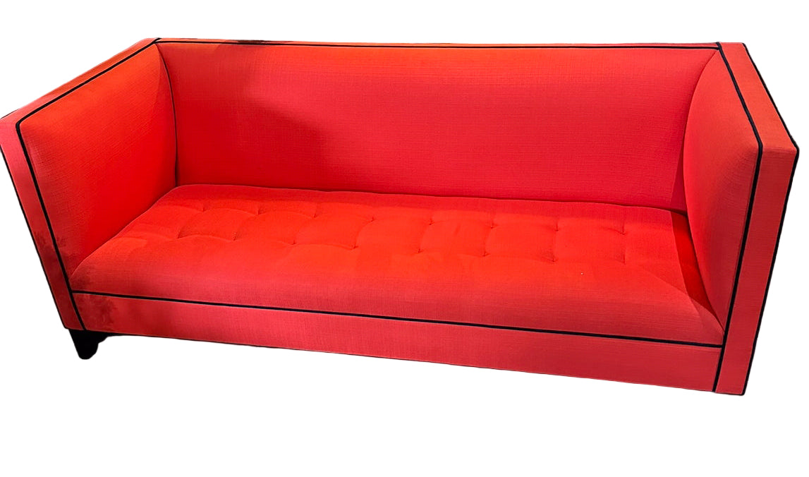 Bright red modern couch with black trim, featuring tufted upholstery. Set in a stylish interior with decorative plants and lamps in the background.