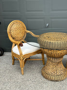 A rattan chair with a woven backrest and white cushion sits beside a round, woven table with a glass top, set against a gray garage door.