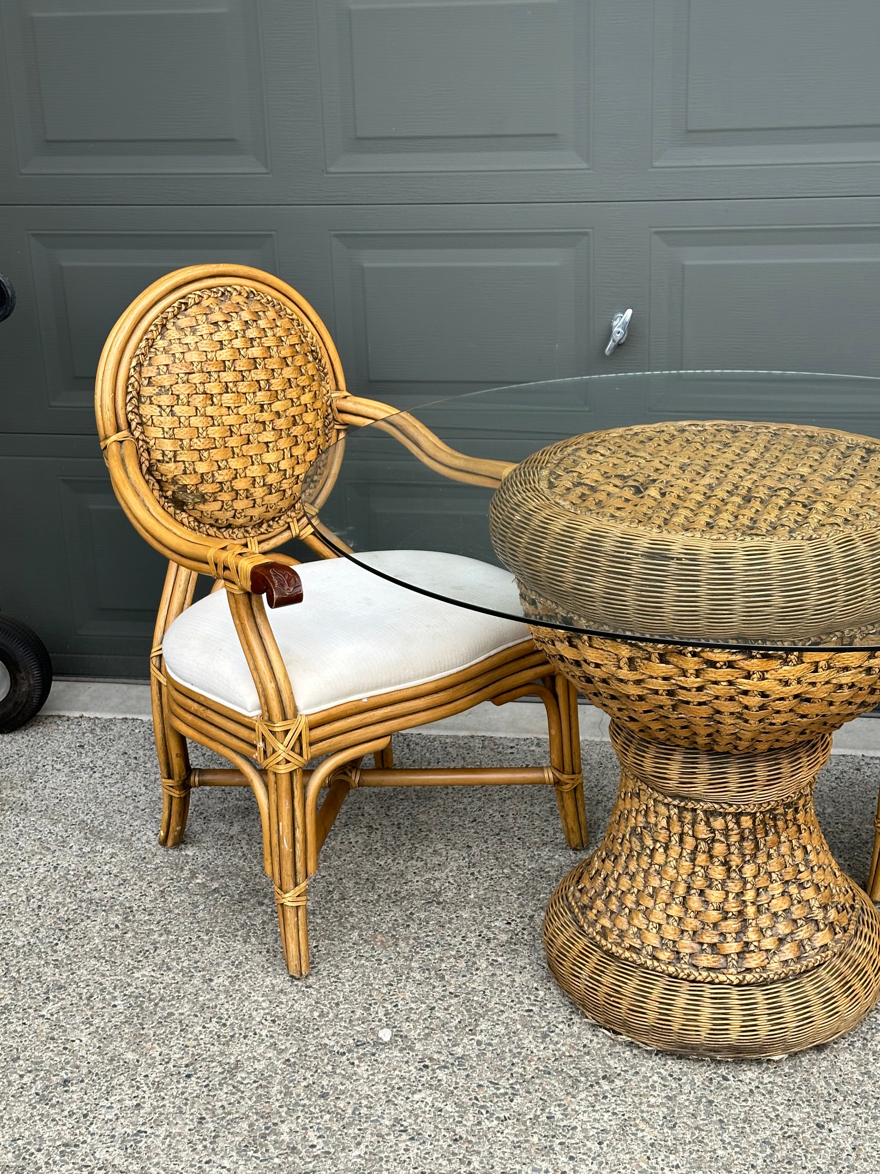 A rattan chair with a woven backrest and white cushion sits beside a round, woven table with a glass top, set against a gray garage door.