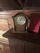 A wooden mantel clock displays the time with a classic design. It rests on a dark surface, with a backdrop of textured wood and a red book.