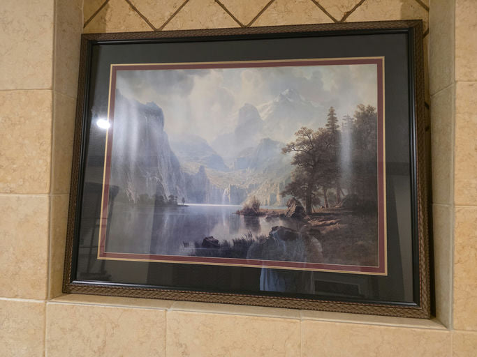 Framed landscape painting featuring mountains, a lake, and lush trees, displayed on a tiled wall. Soft colors evoke a serene atmosphere.