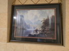 Framed landscape painting featuring mountains, a lake, and lush trees, displayed on a tiled wall. Soft colors evoke a serene atmosphere.