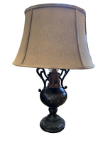 A decorative lamp with a black base and elegant cord design, topped with a beige fabric shade, sits on a patterned rug next to a bed.