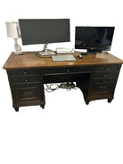 A wooden desk with two monitors, a laptop, and a lamp. The setup includes visible cables and a nightstand with drawers. The floor features laminate planks.