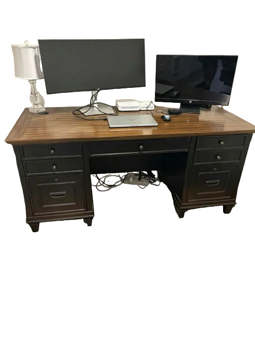 A wooden desk with two monitors, a laptop, and a lamp. The setup includes visible cables and a nightstand with drawers. The floor features laminate planks.