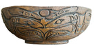 A carved brown wooden bowl features intricate designs, including stylized eyes and geometric patterns, resting on a dark tray with other items.