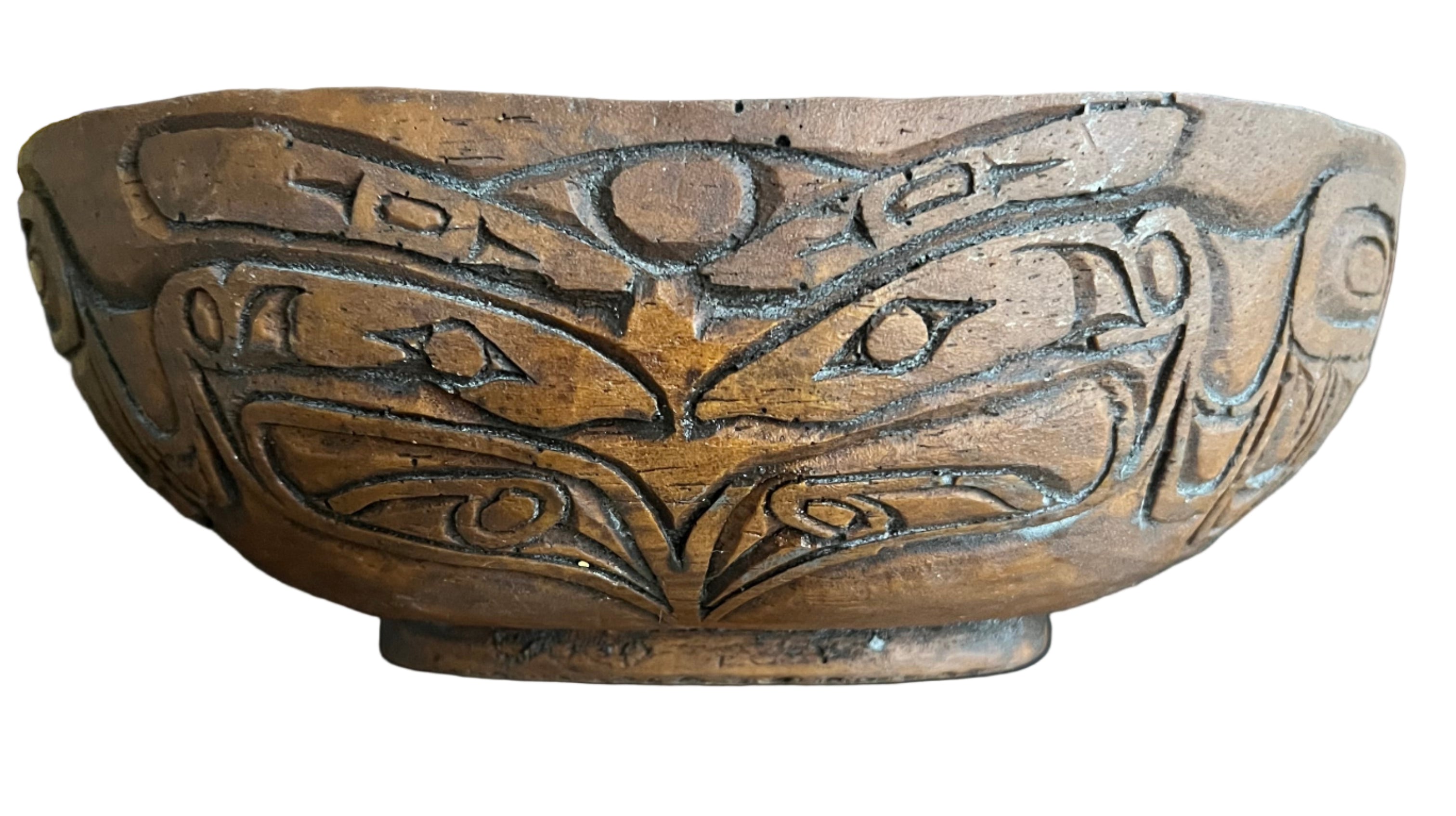 A carved brown wooden bowl features intricate designs, including stylized eyes and geometric patterns, resting on a dark tray with other items.