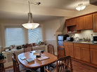 A cozy kitchen with wooden cabinets, a round table, and a pendant light. Photos adorn the windowsill, and kitchen appliances are neatly arranged.