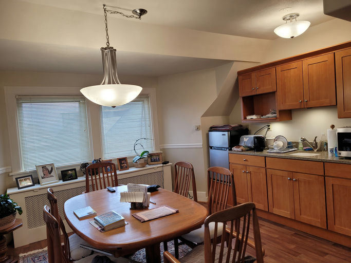 A cozy kitchen with wooden cabinets, a round table, and a pendant light. Photos adorn the windowsill, and kitchen appliances are neatly arranged.