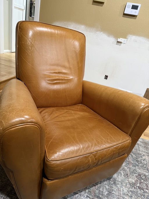 A worn, tan leather armchair with visible creases and scratches, set against a partially painted wall and hardwood flooring, typical of indoor furnishings.