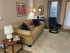 Cozy living room with a beige sofa, colorful pillows, a lamp, and a wooden table. A navy rocking chair and books are nearby, with curtains letting in light.