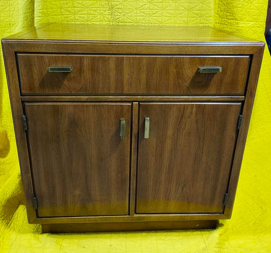 A wooden cabinet with two doors and a drawer, featuring metallic handles. The background is bright yellow, highlighting the furniture piece.