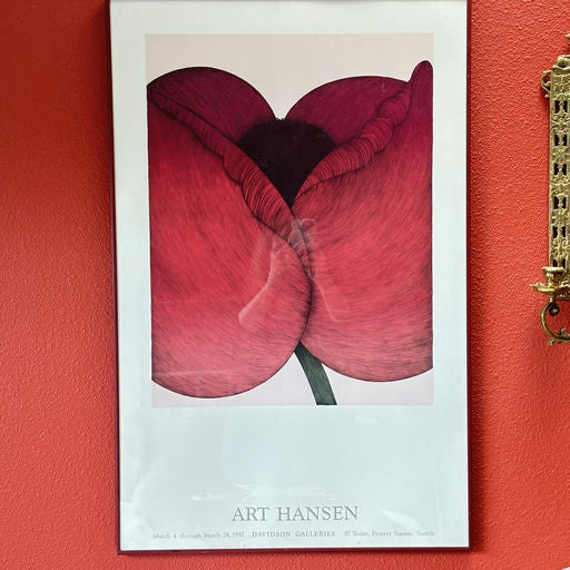 A framed art print featuring two large red flowers with intricate details, displayed against a vibrant red wall. Information about the artist is included.