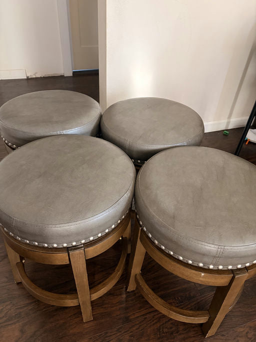 Four round stools with gray upholstery and decorative silver studs stacked closely together on a wooden floor in a well-lit room.