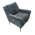 A modern gray velvet armchair with gold metal legs, positioned in a showroom setting on a patterned rug in front of a brown sectional sofa.