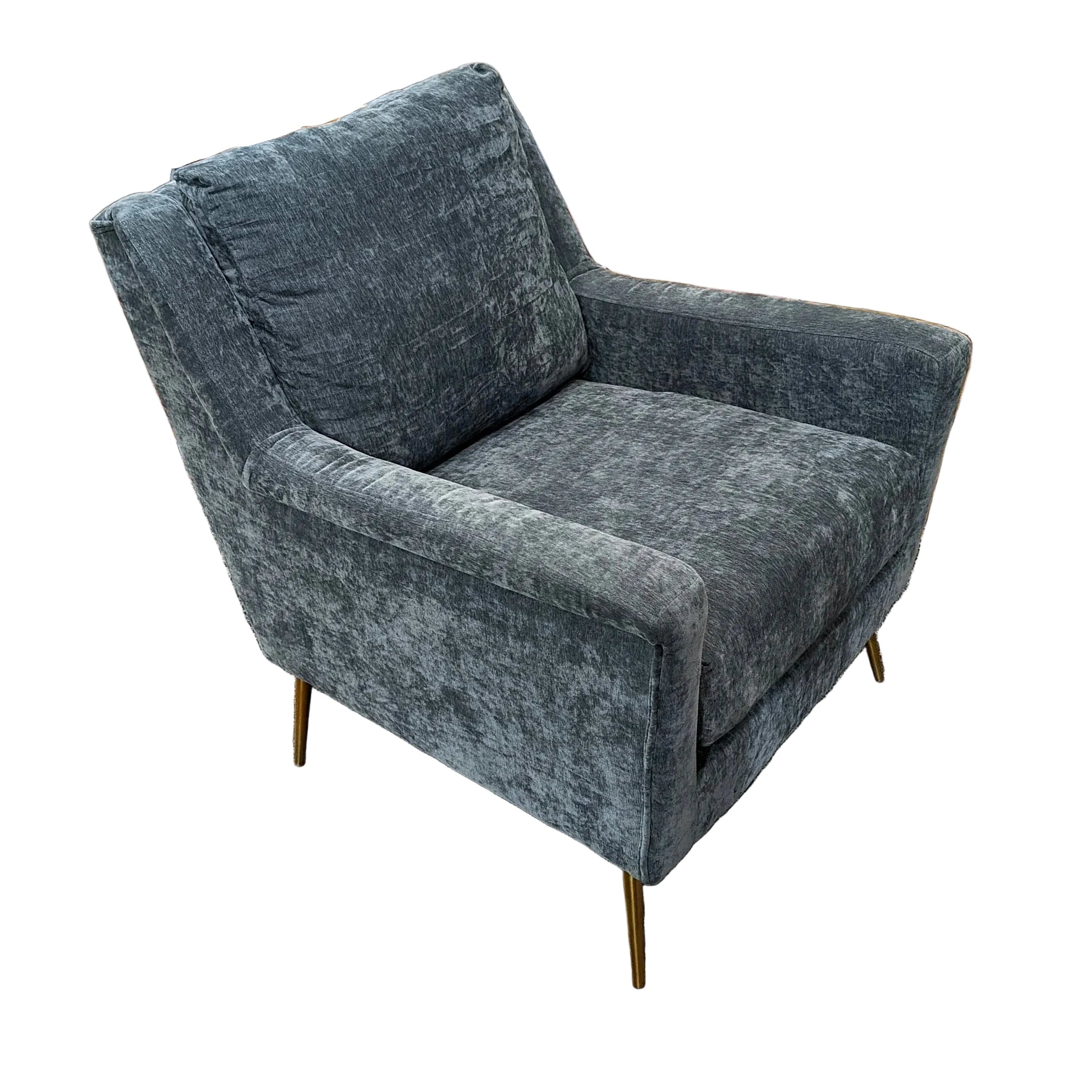 A modern gray velvet armchair with gold metal legs, positioned in a showroom setting on a patterned rug in front of a brown sectional sofa.