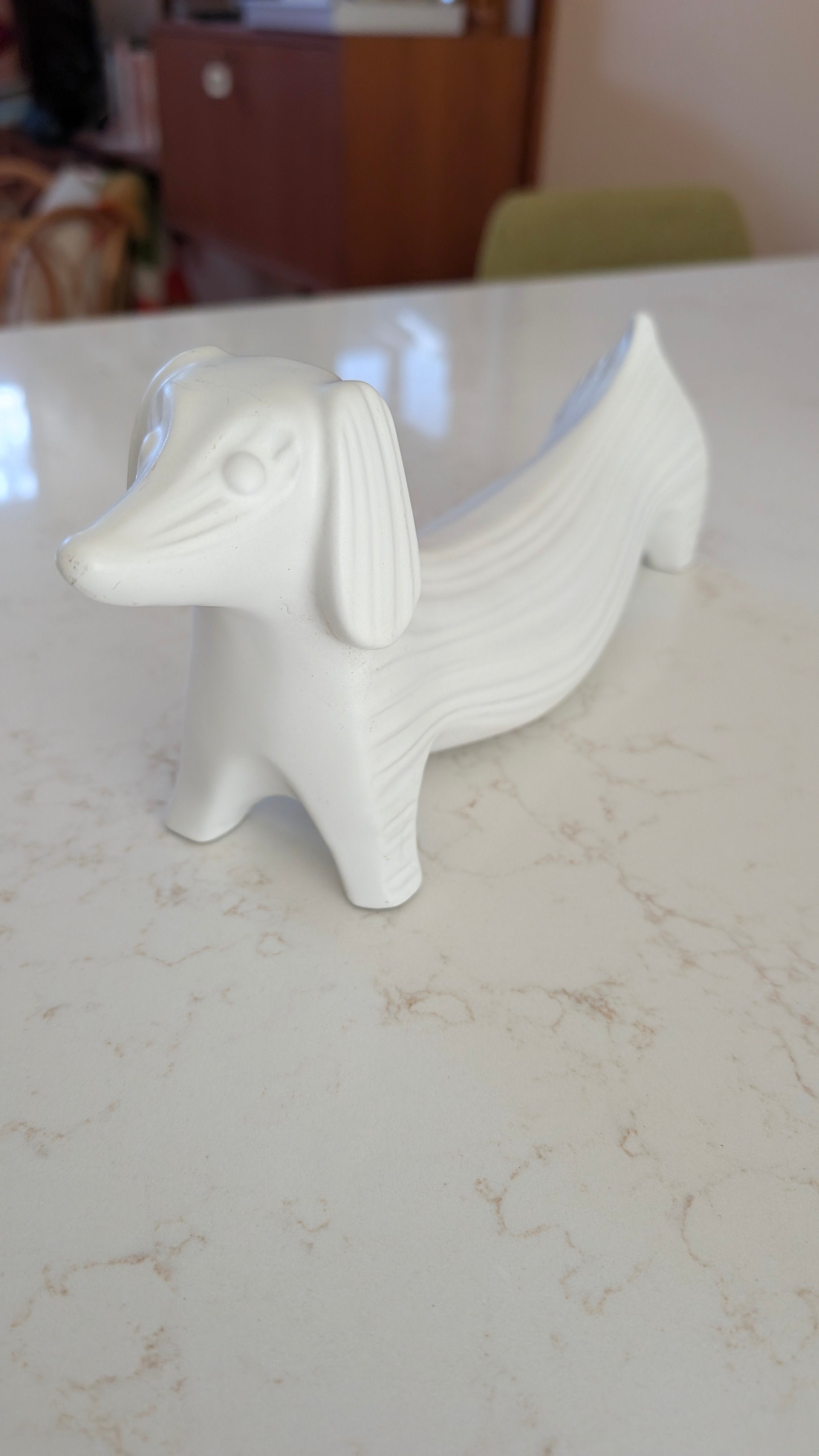 A white ceramic figurine shaped like a dachshund, with textured lines, placed on a smooth marble surface. Background includes blurred furniture.