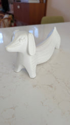 A white ceramic figurine shaped like a dachshund, with textured lines, placed on a smooth marble surface. Background includes blurred furniture.