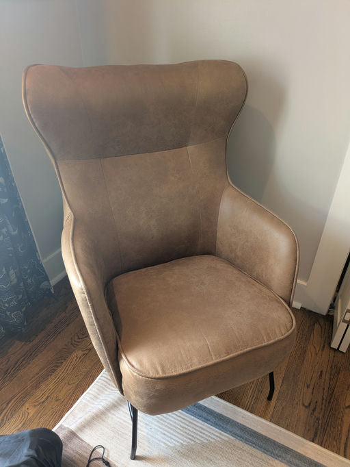 A stylish, modern armchair featuring a light brown faux leather finish and sleek black legs, positioned on a patterned rug against a wall.